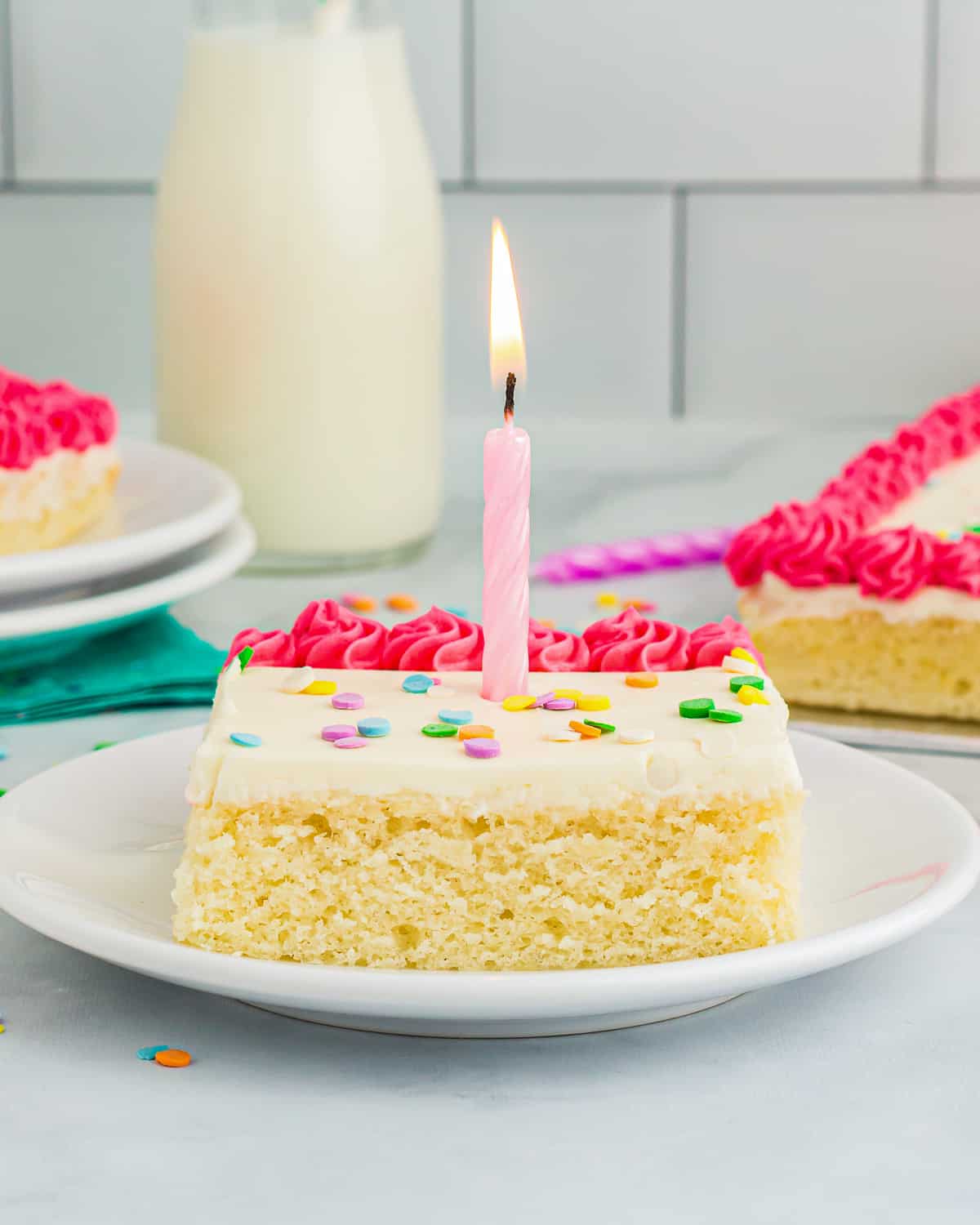 side view of a slice of vanilla sheet cake on a white plate with a lit pink birthday candle on top.