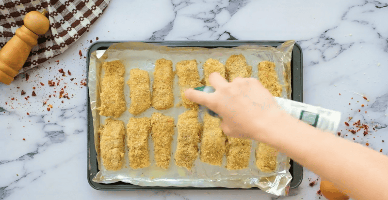 spraying olive oil on fish sticks before baking