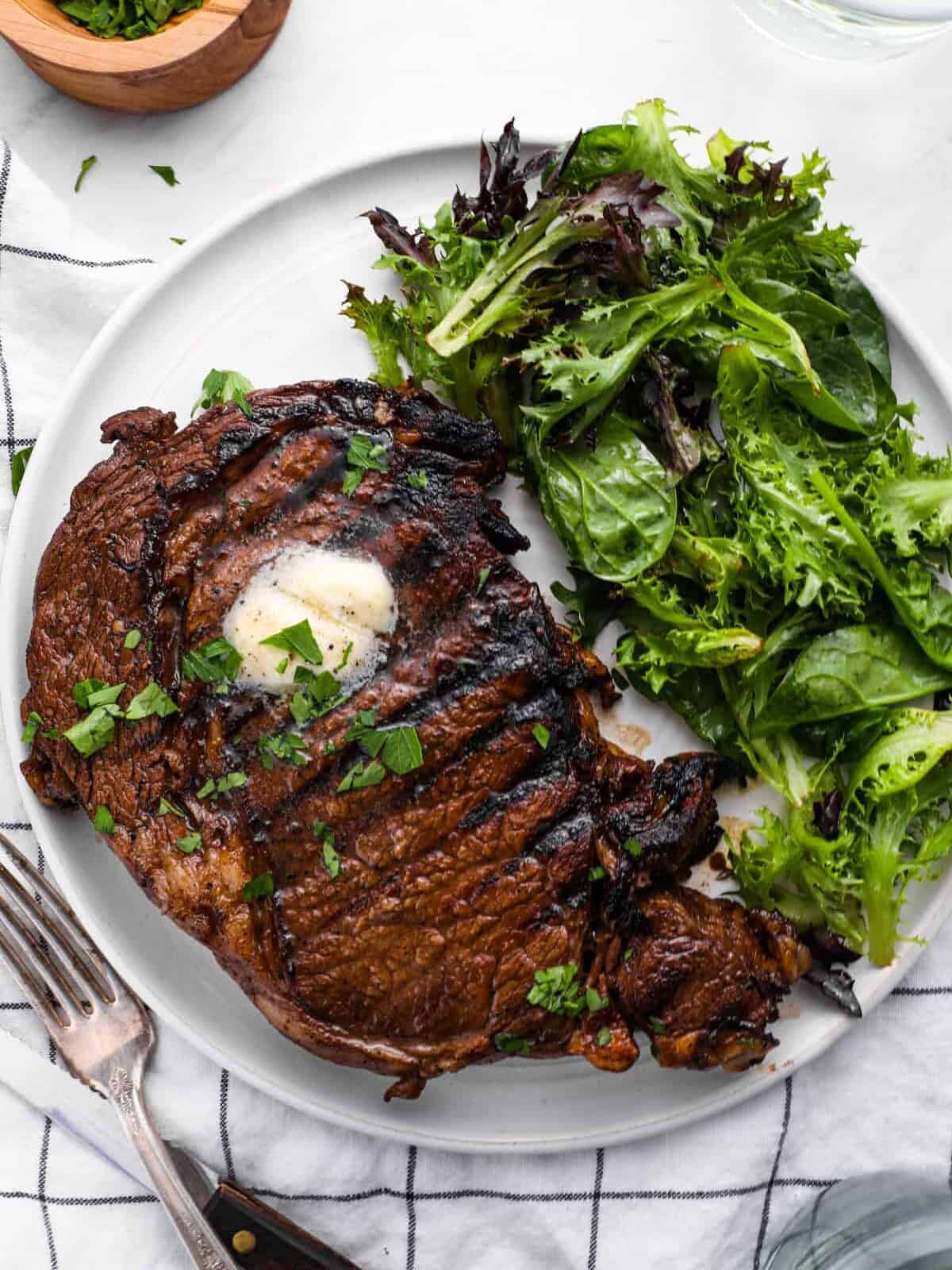 overhead view of a marinated ribeye steak on a white plate with butter and salad greens.