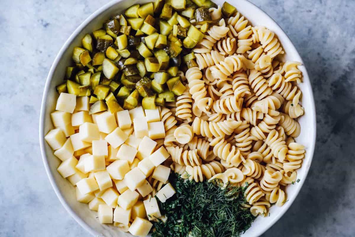 ingredients for dill pickle pasta salad (cubes of cheese, chopped pickles, dill, and rotini pasta) in a white bowl.