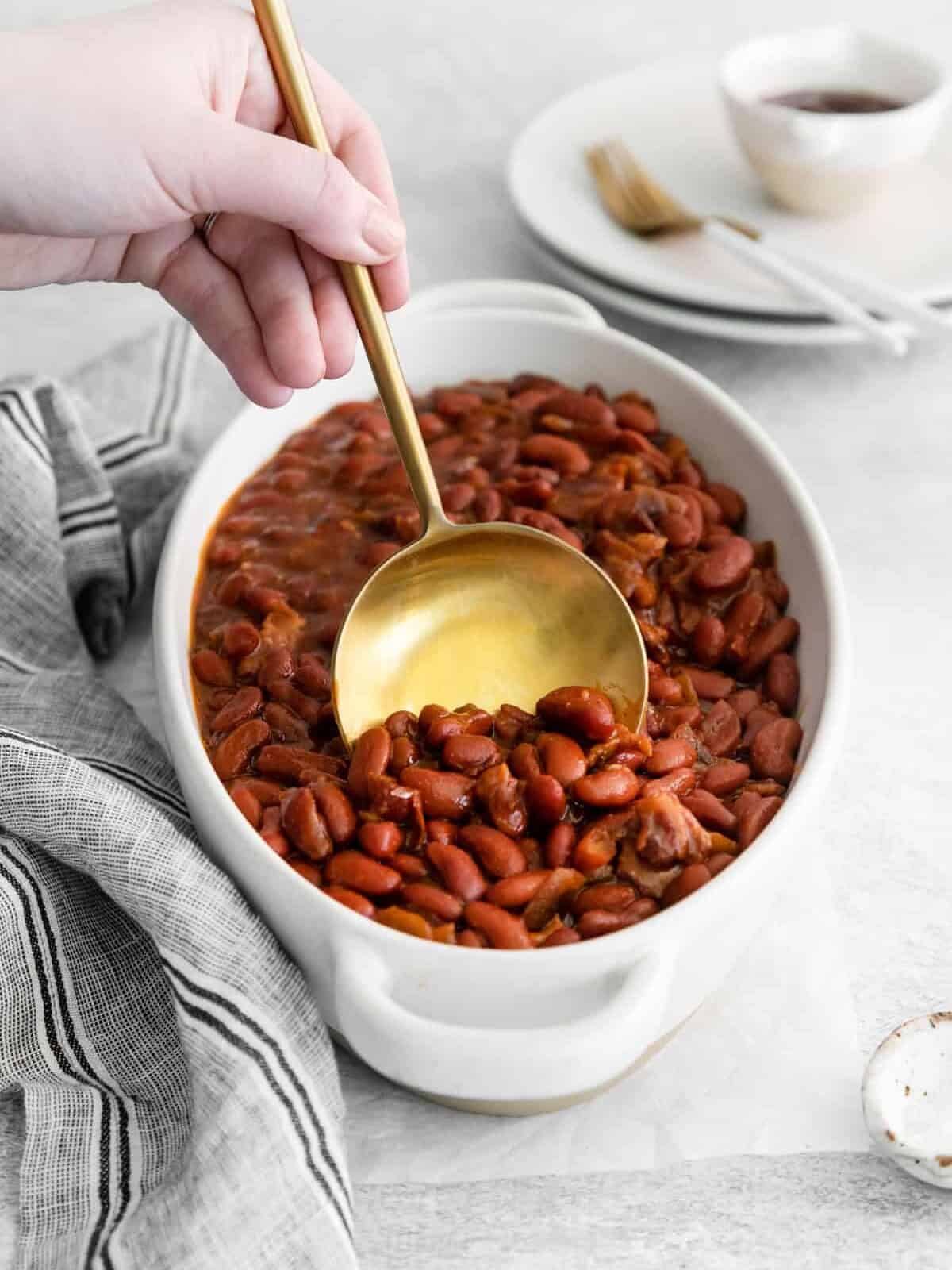 a hand using a spoon to scoop instant pot bbq baked beans from a white serving dish.