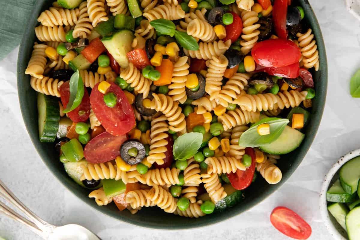 partial overhead view of pasta salad filled with colorful vegetables (tomatoes, corn, olives, pears, squash, and more) in a gray bowl.