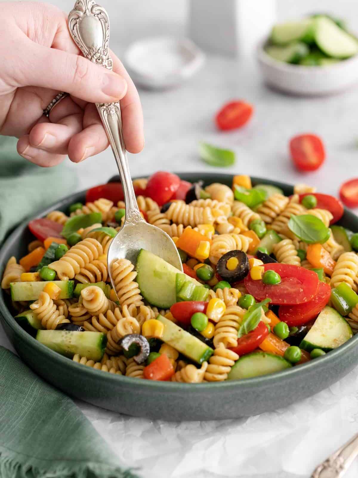 three-quarters view of a hand scooping veggie rainbow pasta salad from a gray bowl with a spoon.