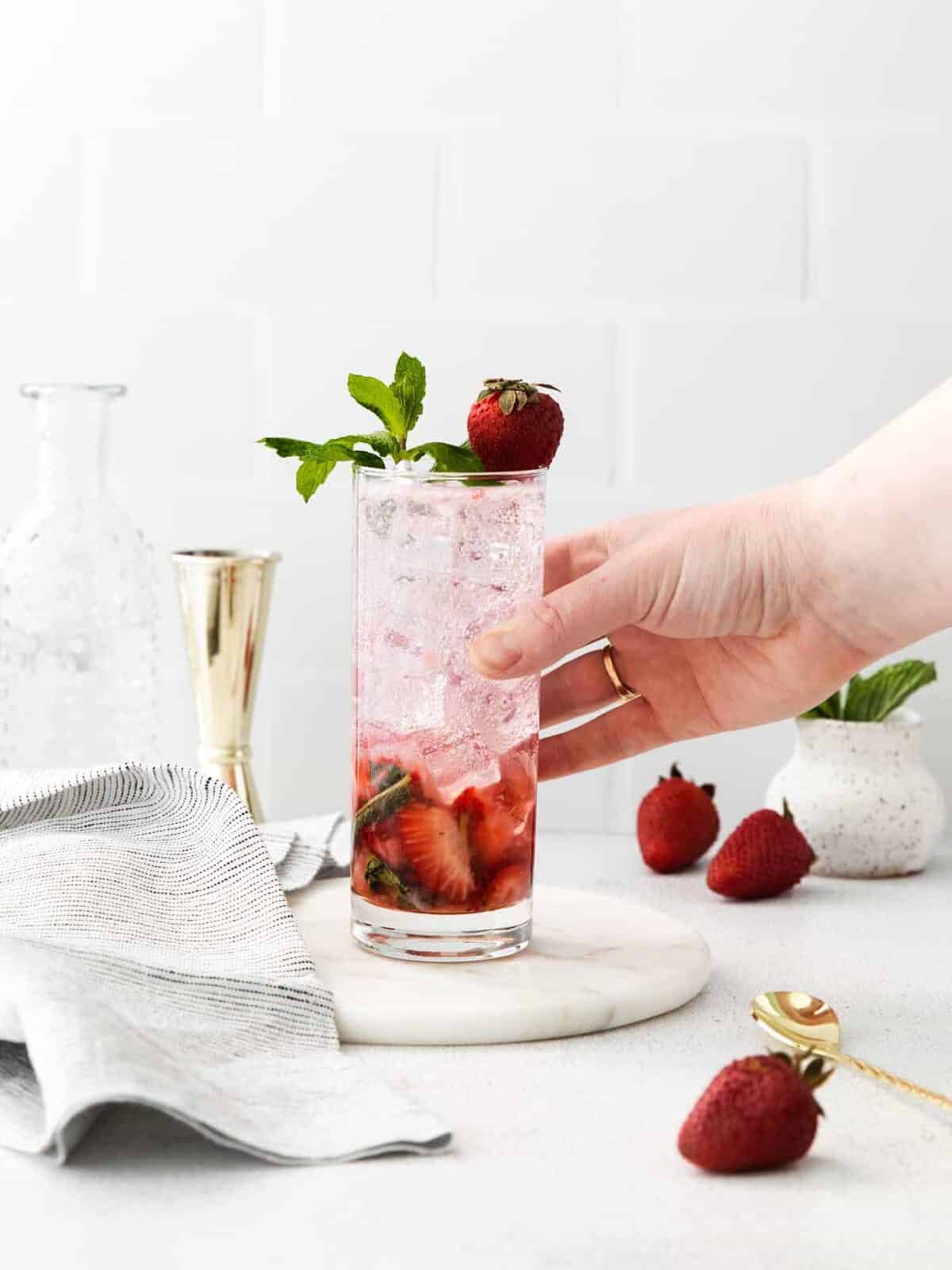 side view of a hand grabbing a strawberry mojito in a highball glass garnished with mint sprigs and a strawberry.