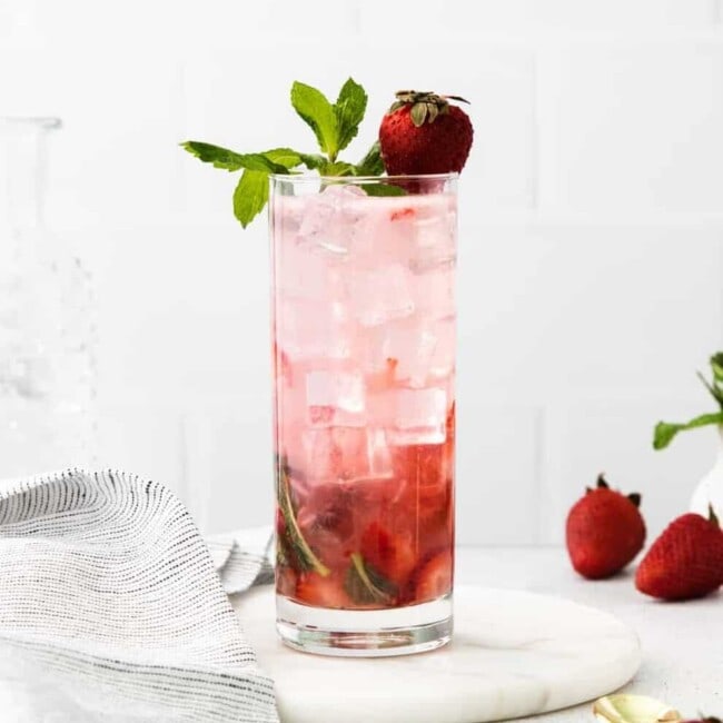 side view of a strawberry mojito in a highball glass on a white plate topped with mint sprigs and a strawberry.