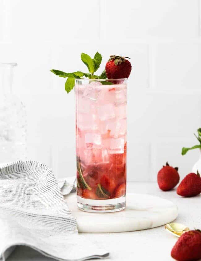 side view of a strawberry mojito in a highball glass on a white plate topped with mint sprigs and a strawberry.