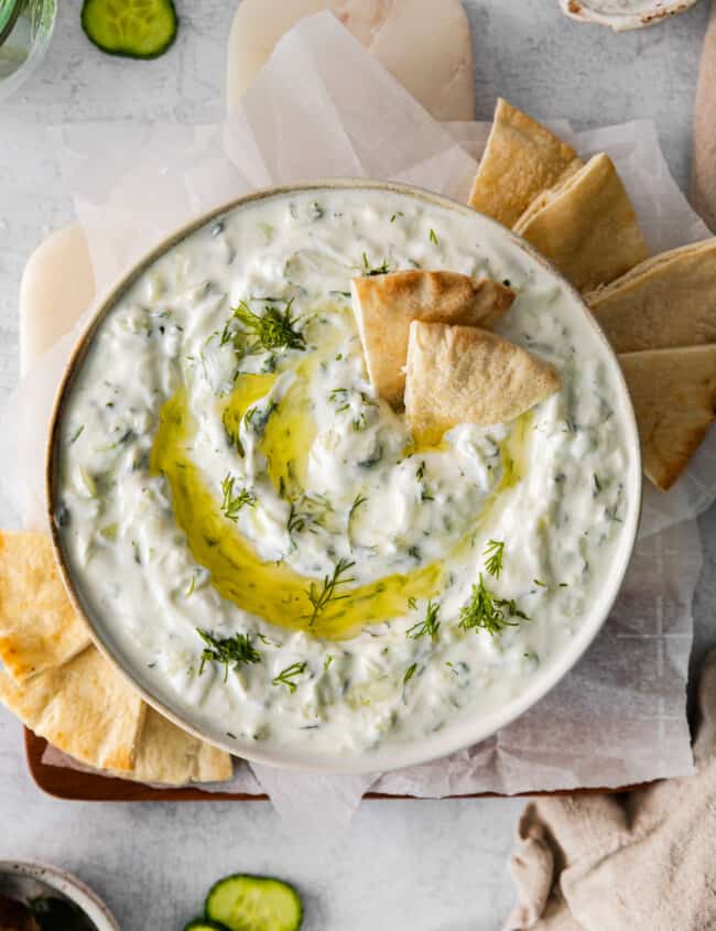 overhead view of tzatziki dip in a white bowl with a drizzle of olive oil and pita chips.