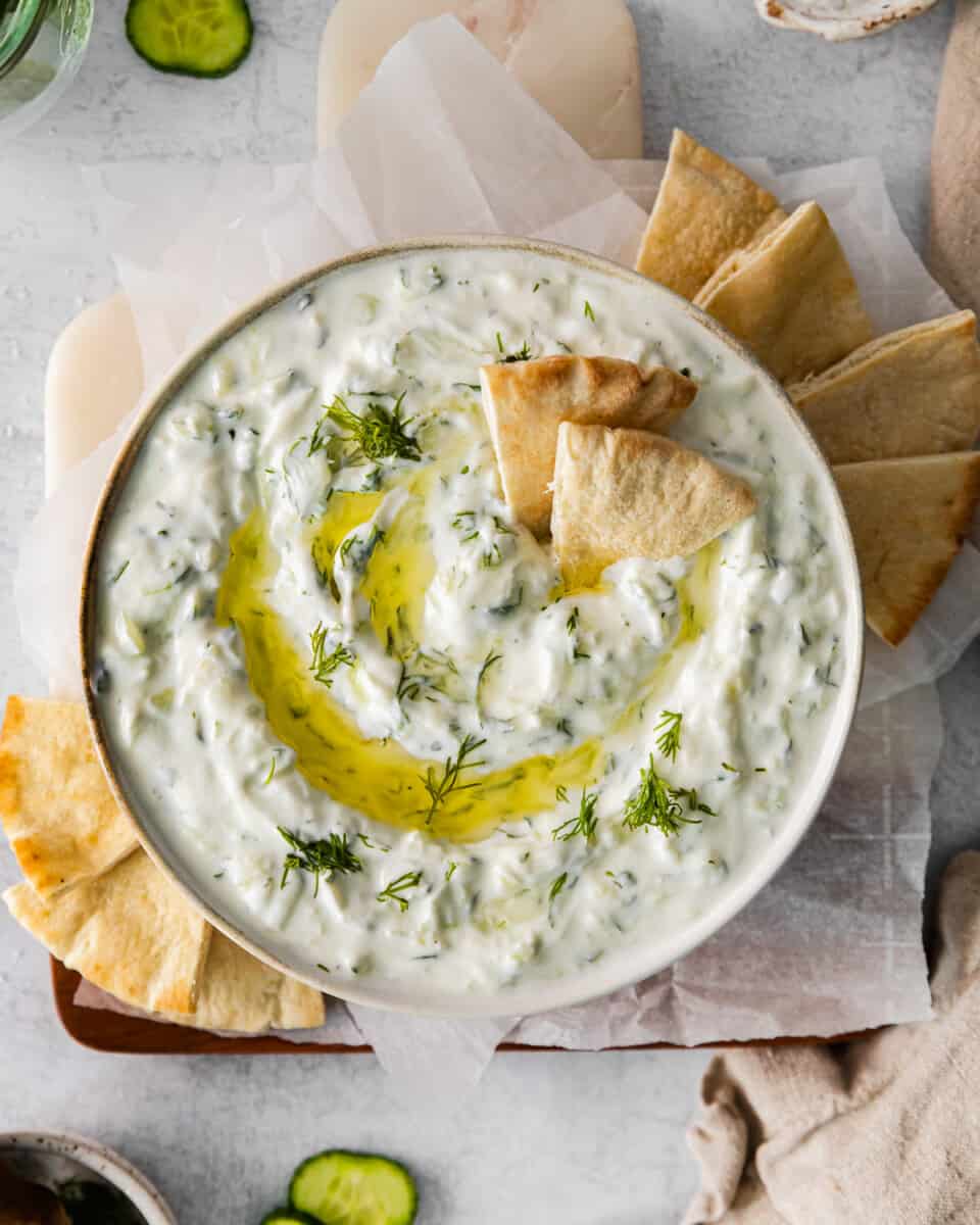 overhead view of tzatziki dip in a white bowl with a drizzle of olive oil and pita chips.