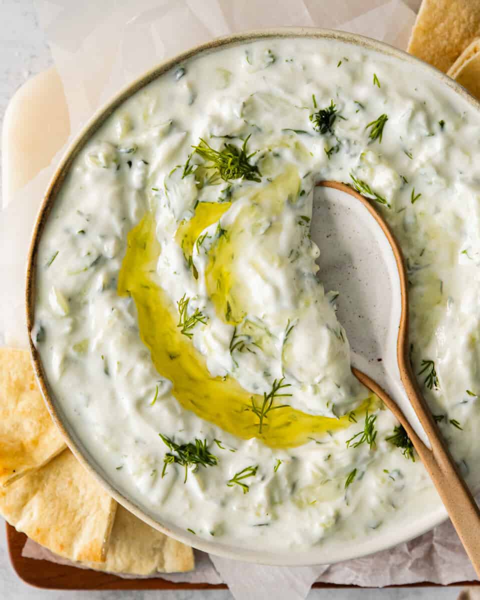 close up overhead view of tzatziki dip in a white bowl with a drizzle of olive oil and a wooden spoon.