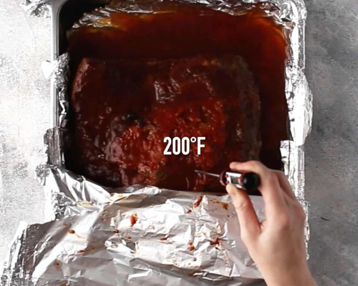 Cooked oven-baked brisket in foil-lined pan being checked with thermometer at 200°F