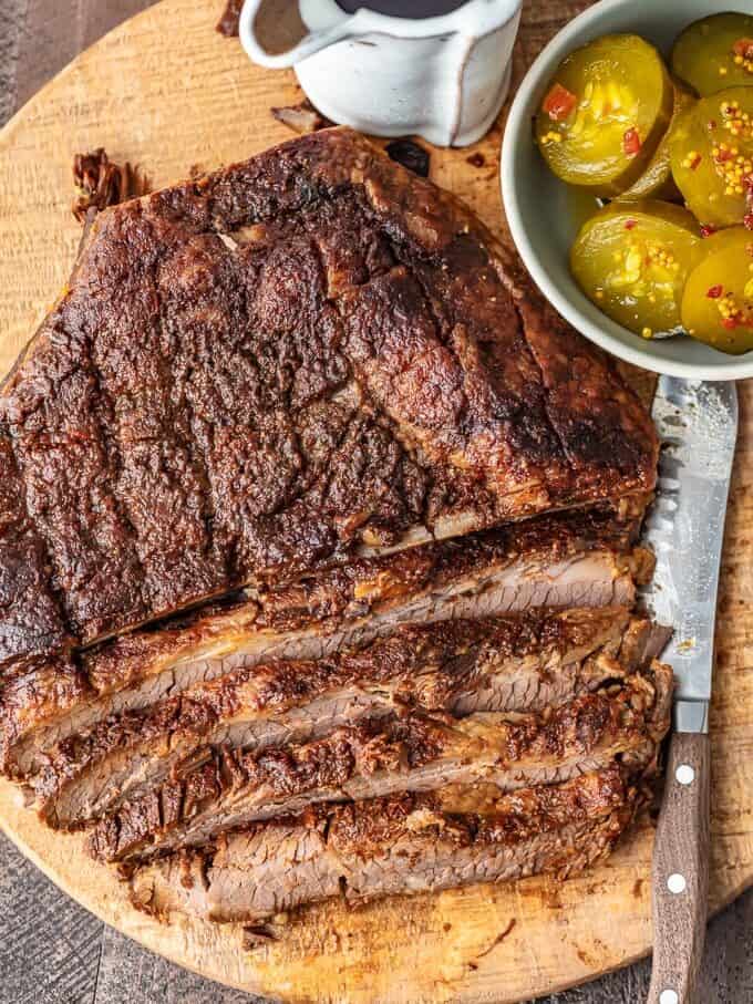 overhead view of baked beef brisket on a cutting board, with a cup of BBQ sauce and a bowl of pickles set to the side