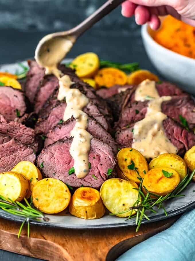 Beef Tenderloin is one of the best cuts of meat, so it deserves to be cooked right. This beef tenderloin roast is perfectly cooked and full of flavor. Learn how to cook beef tenderloin for an amazing main course dish to serve on the holidays or special occasions. This is the BEST beef tenderloin recipe, just wait and see! drizzling brand peppercorn sauce over beef tenderloin