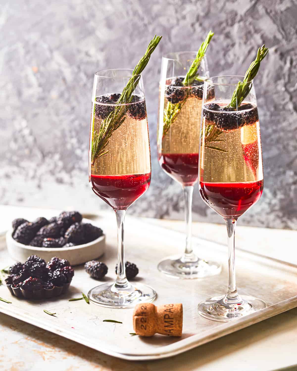 side view of 3 blackberry champagne cocktails on a metal tray with a bowl of blackberries and a champagne cork.