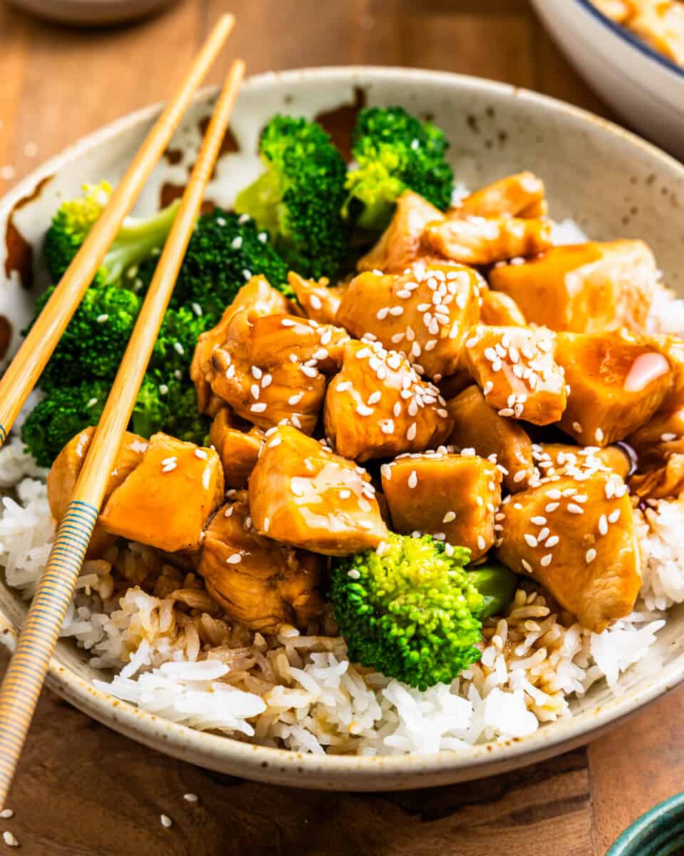 three-quarters view of teriyaki chicken in a white bowl with rice and broccoli and chopsticks.