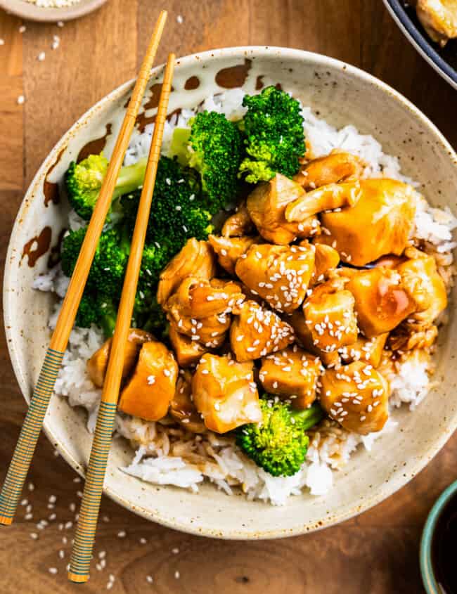 overhead view of teriyaki chicken in a white bowl with rice and broccoli and chopsticks.