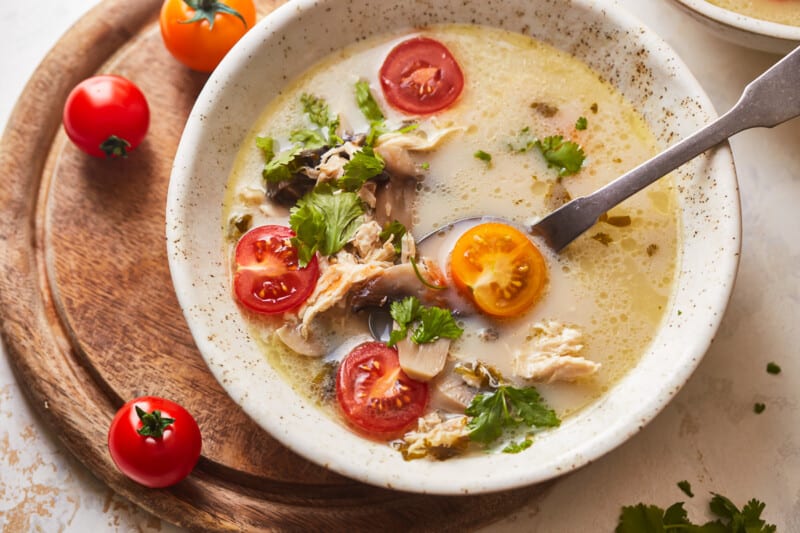 a serving of Thai coconut chicken soup in a white bowl with a spoon on top of a wooden cutting board.