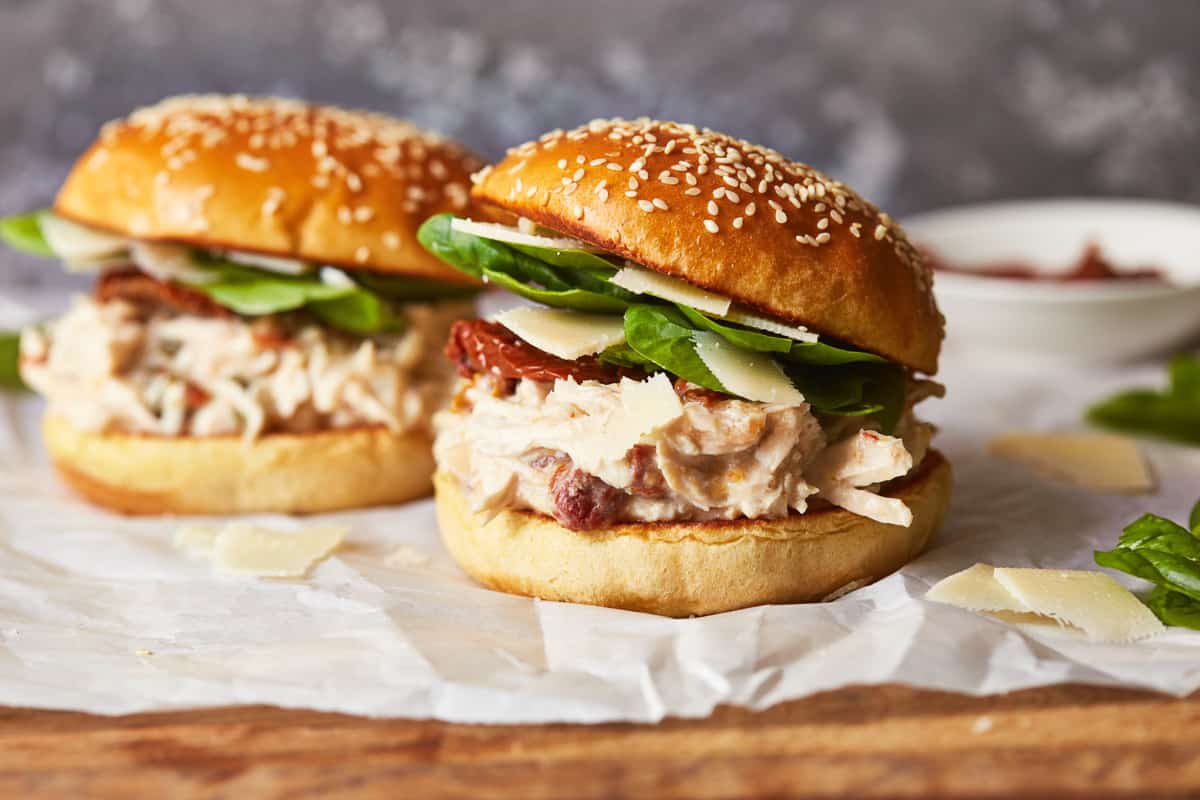 side view of 2 chicken alfredo sandwiches on parchment paper on a wooden cutting board.