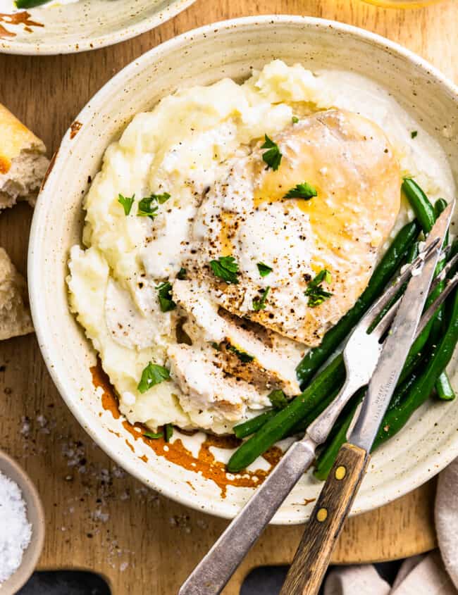 close-up overhead view of crockpot ranch chicken on a white plate with green beans, mashed potatoes, and a fork and knife.