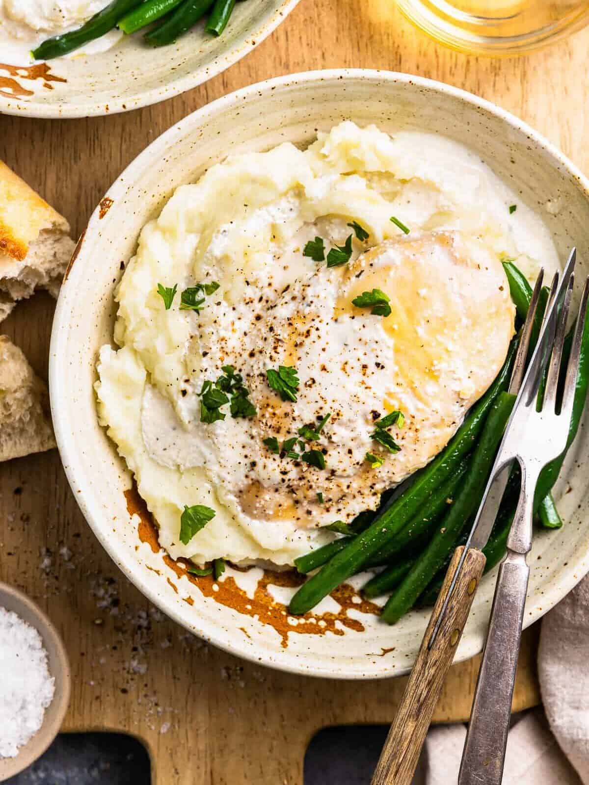 overhead view of crockpot ranch chicken on a white plate with green beans, mashed potatoes, and a fork and knife.