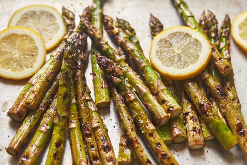 three-quarters view of grilled asparagus on a baking sheet with lemon wheels.