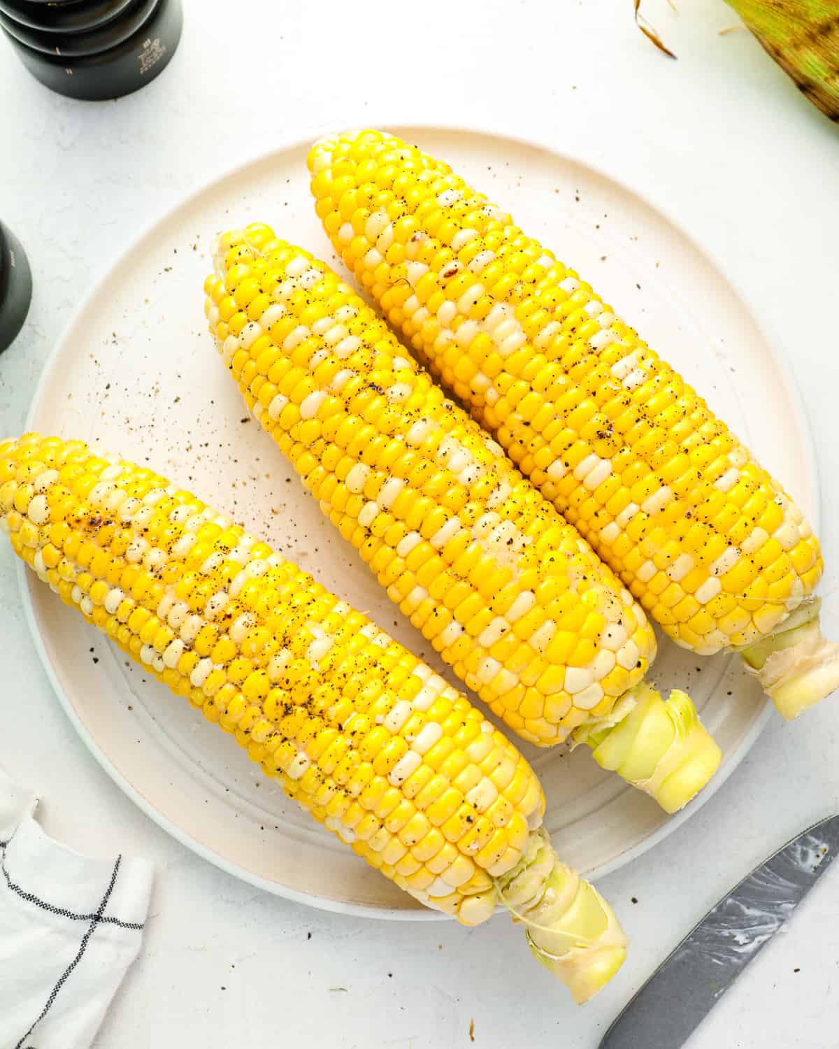 overhead view of 3 grilled corn on the cobs on a white plate seasoned with salt and pepper.