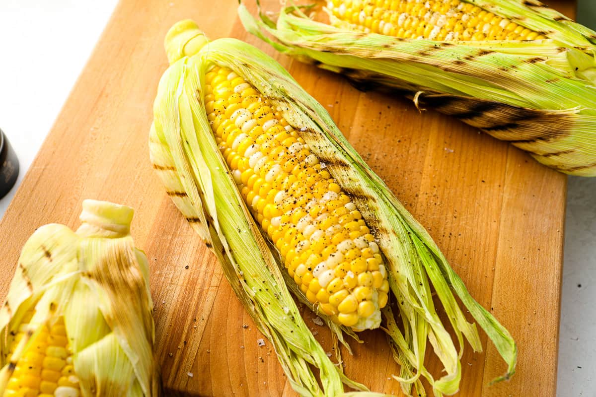 three-quarters view of grilled corn on the cob with the husk peeled back on a wooden cutting board.