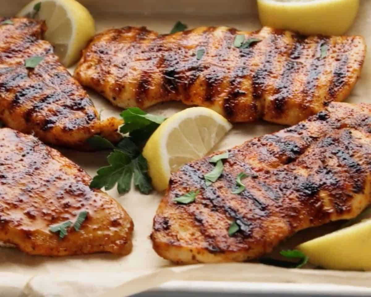 Grilled marinated chicken breasts with char marks served with lemon wedges and fresh parsley on a platter.