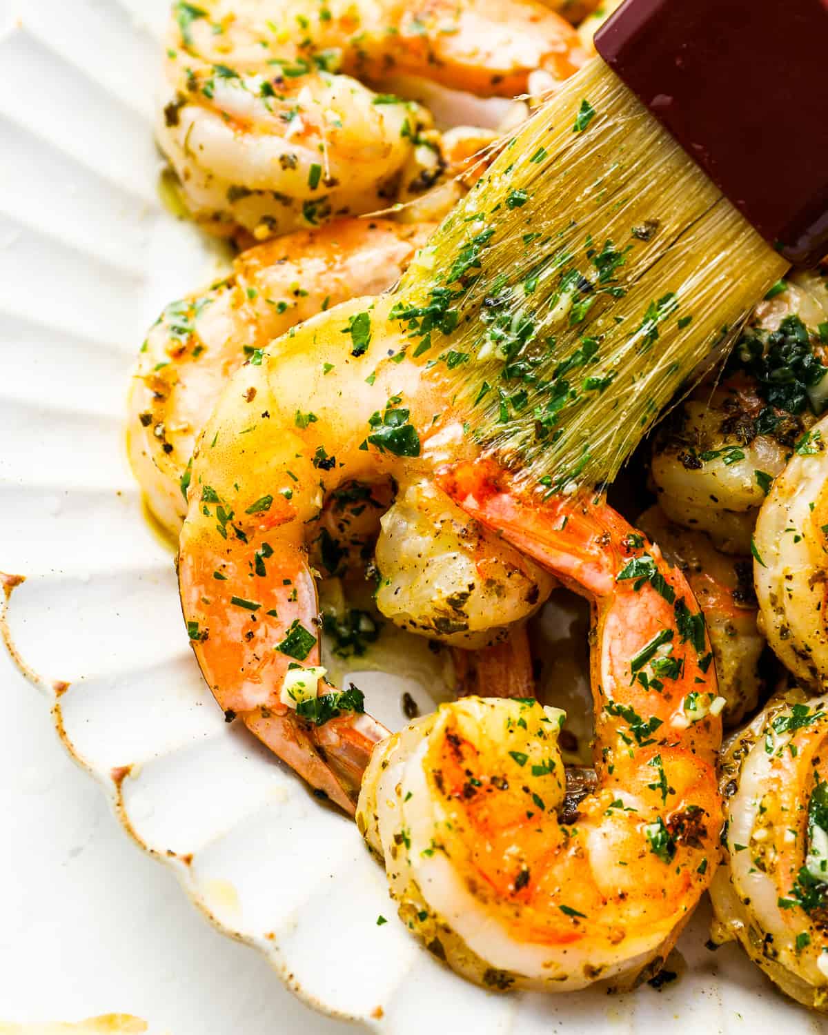 a pastry brush brushing lemon garlic butter over grilled shrimp on a white plate.