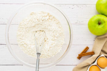 a bowl of flour, eggs and apples on a wooden table.