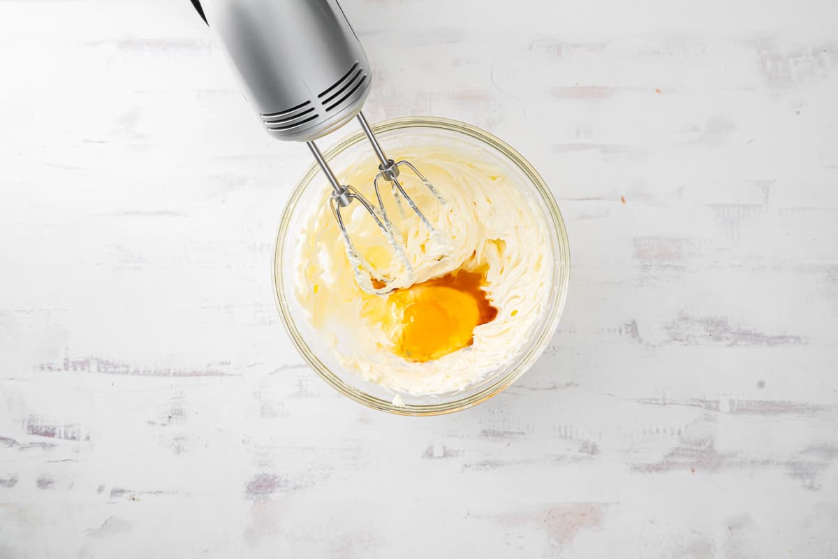 egg and vanilla added to whipped cream cheese in a glass bowl with a hand mixer.