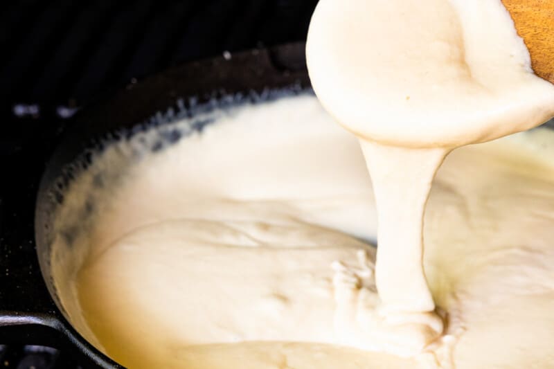 a wooden spoon lifting cheese fondue from a pot to show the viscosity.