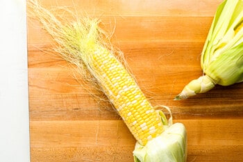 a corn con with the corn silk still attached on a wooden cutting board.
