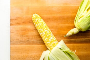 a corn cob with the husk peeled back and the silks removed on a wooden cutting board.