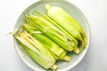 4 ears of corn with the husks folded over them in a green bowl filled with salty water.