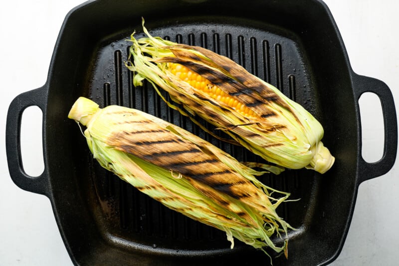 2 grilled ears of corn in the husks on a grill pan.