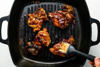 a pastry brush brushing huli huli sauce over 4 grilled chicken thighs in a grill pan.