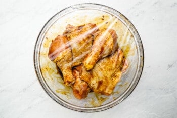 chicken thighs and drumsticks marinating in korean bbq sauce in a glass bowl with plastic wrap over top.