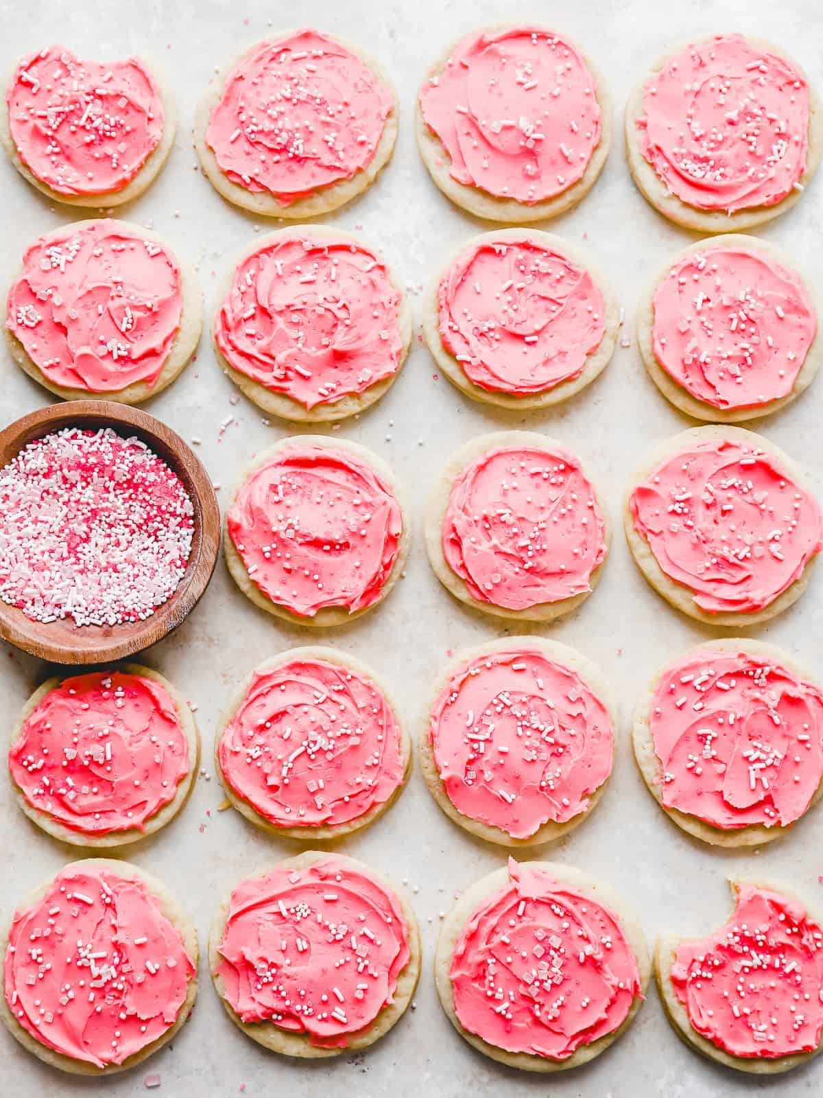 overhead view of 19 pink frosted lofthouse cookies in a grid pattern with a bowl of sprinkles.