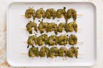 overhead view of 20 raw shrimps threaded on skewers and coated in pesto on a baking sheet.