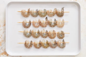 20 raw shrimps threaded on skewers on a baking sheet.