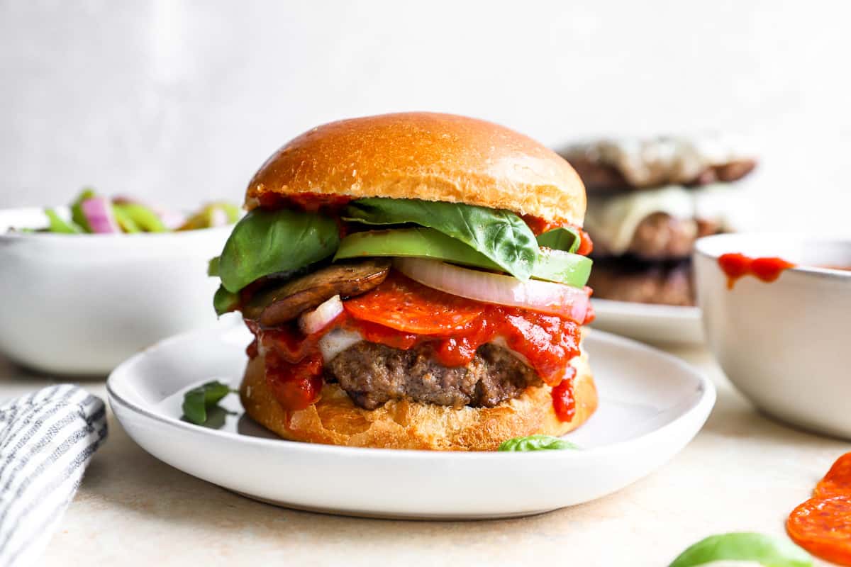 a burger on a plate filled with pizza toppings, including tomatoes, mushrooms, and basil.