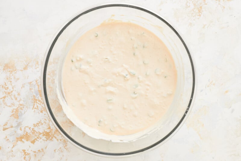 cocktail sauce and green onions added to cream cheese, sour cream, and mayonnaise mixed together in a glass bowl.