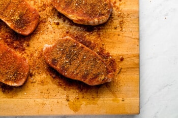 4 seasoned raw pork chops on a wooden cutting board.