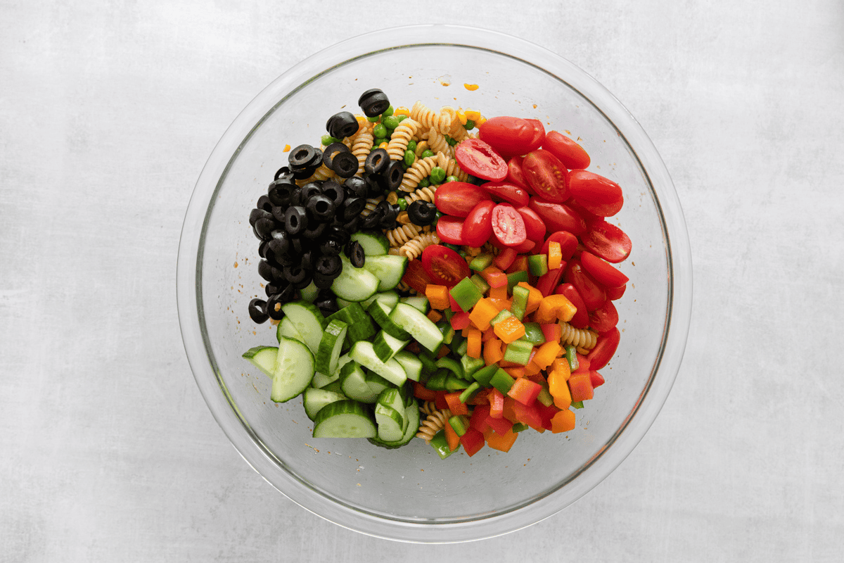 Adding tomatoes, cucumbers, peppers and olives to the bowl with pasta, corn and peas.