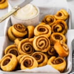 three-quarters view of mini cinnamon rolls in a serving tray with a small pot of frosting and a knife.