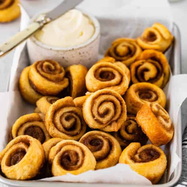 three-quarters view of mini cinnamon rolls in a serving tray with a small pot of frosting and a knife.