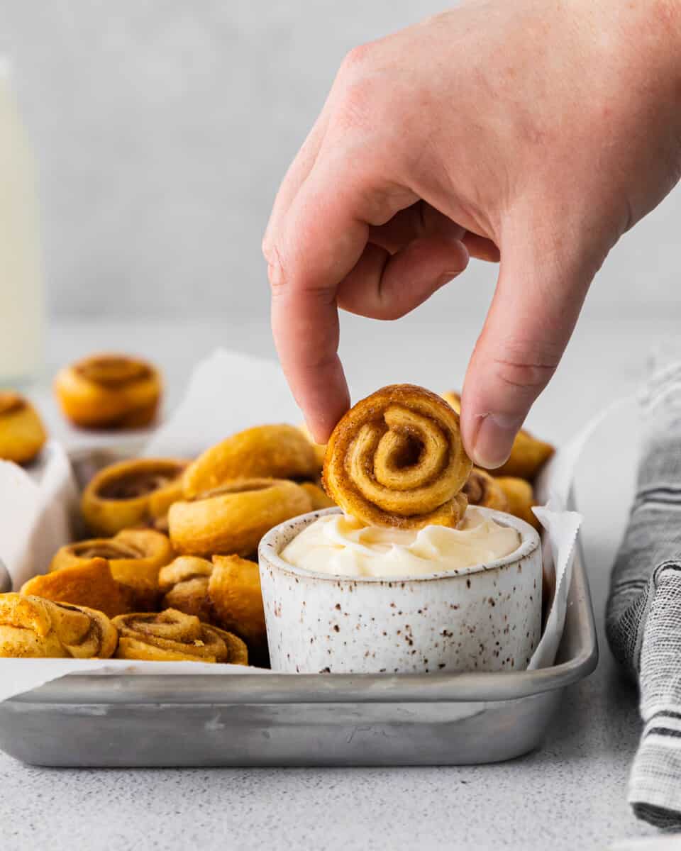 side view of a hand dipping a mini cinnamon roll into a small pot of icing.