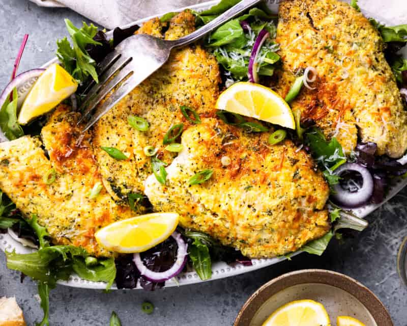 overhead view of parmesan crusted tilapia on an oval platter with salad greens and lemon wedges.