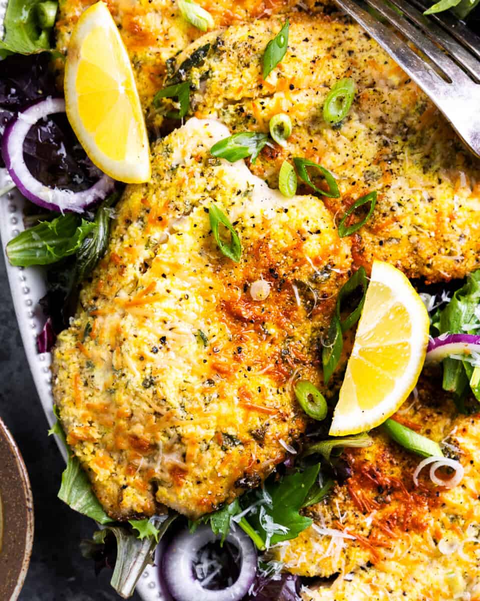 close up view of a parmesan crusted tilapia filet on an oval platter with salad greens and lemon wedges.