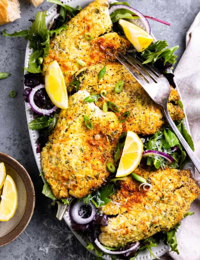 overhead view of parmesan crusted tilapia filets on an oval platter with salad greens, lemon wedges, and a fork.